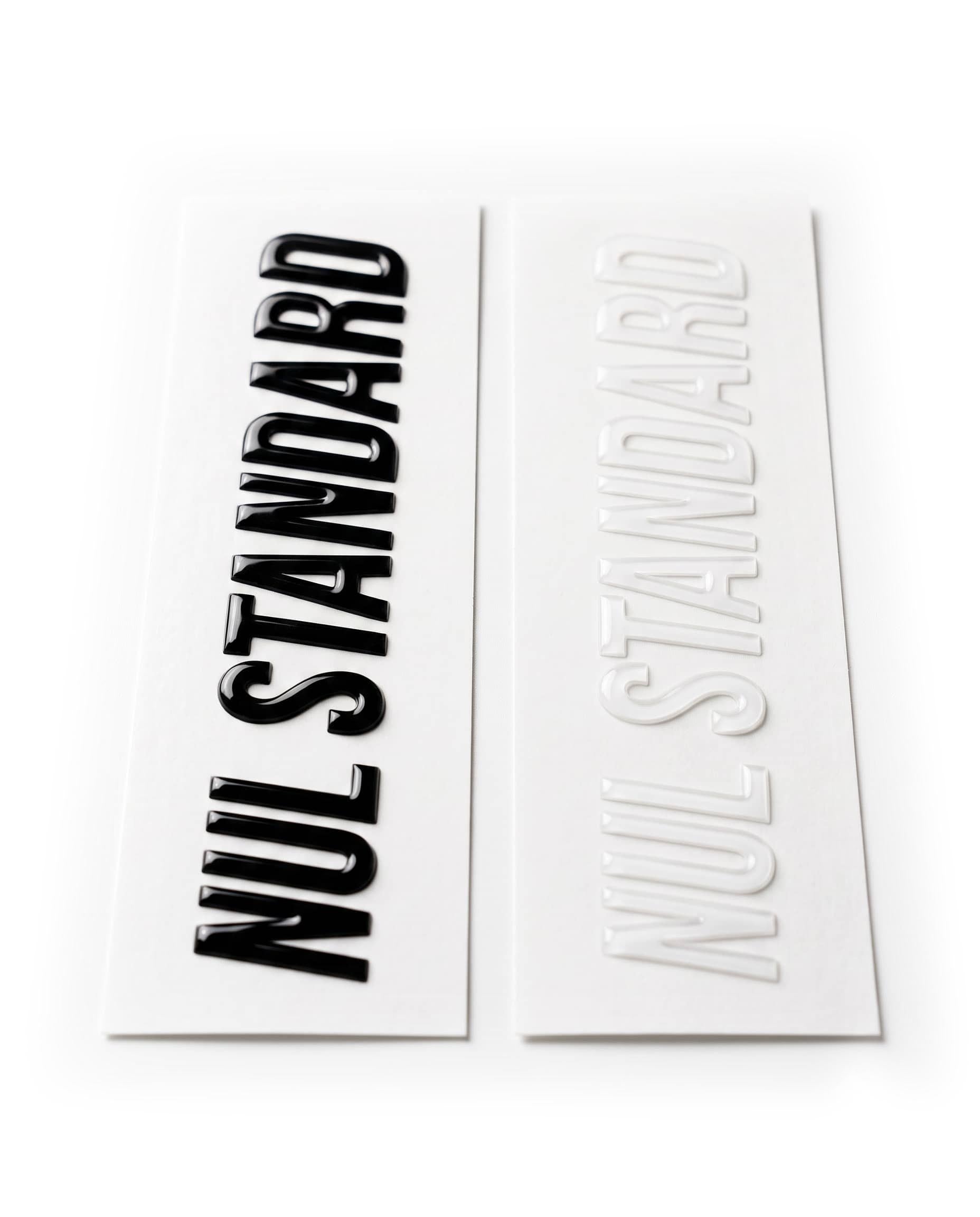 Two rectangular decals or stickers on a white background, one printed with the words 'NUL STANDARD' in bold black and the other in raised white.