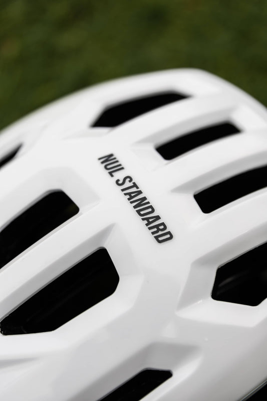 Close-up of a white bicycle helmet with black vent slots and the text 'NUL STANDARD' printed on the side, set against a blurred green background.